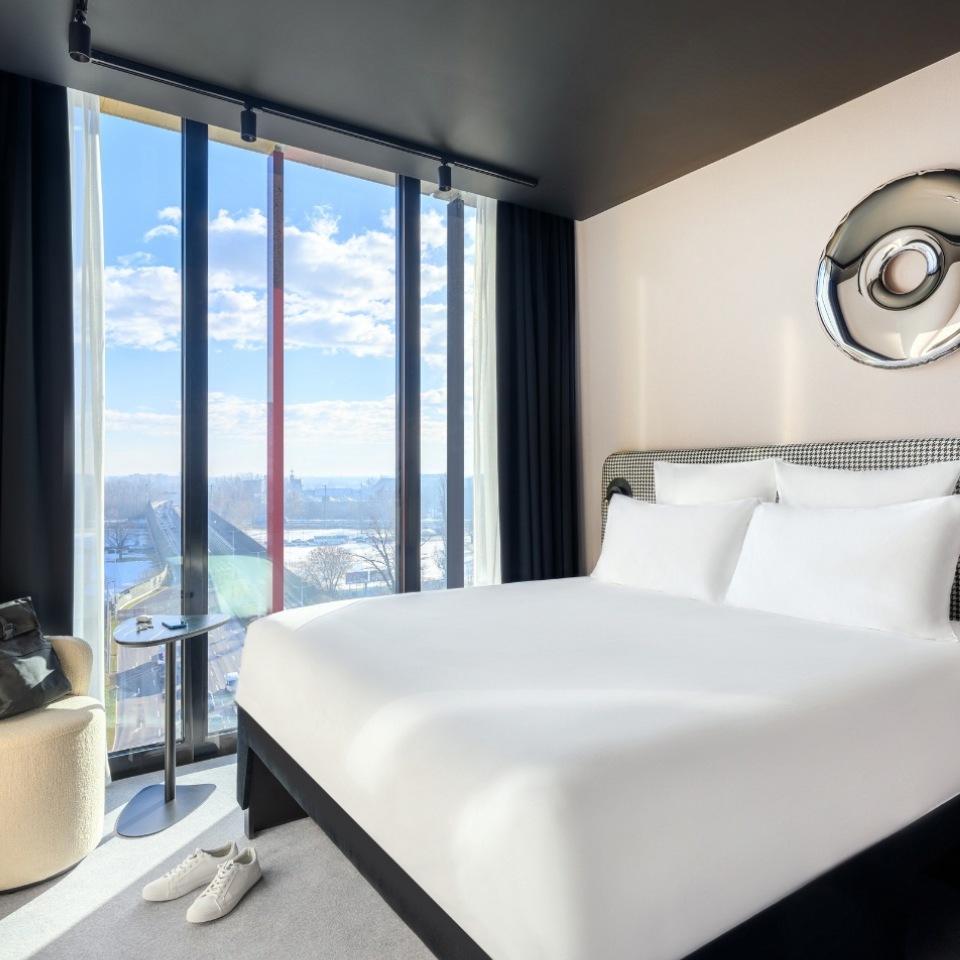 Chambre d’hôtel avec un grand lit, un mobilier élégant et des baies vitrées du sol au plafond offrant une belle vue et une lumière du jour éclatante.