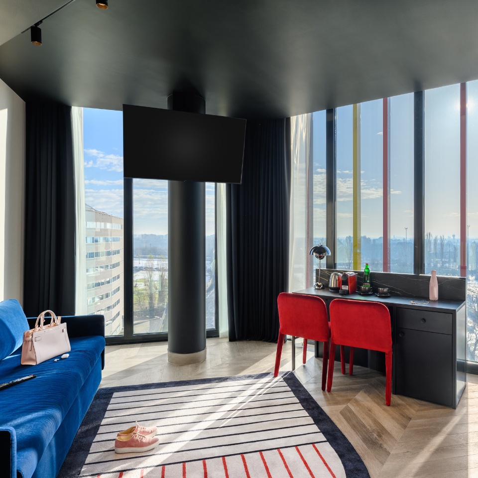 Une chambre d’hôtel élégante avec un canapé bleu, des chaises rouges et un bureau près de grandes fenêtres, avec vue sur les toits de la ville.