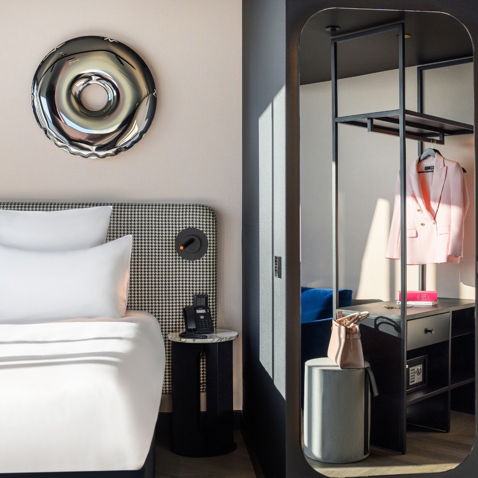 Une chambre d’hôtel moderne avec un lit élégant aux oreillers blancs, une table de chevet épurée et un espace penderie minimaliste avec un blazer rose.