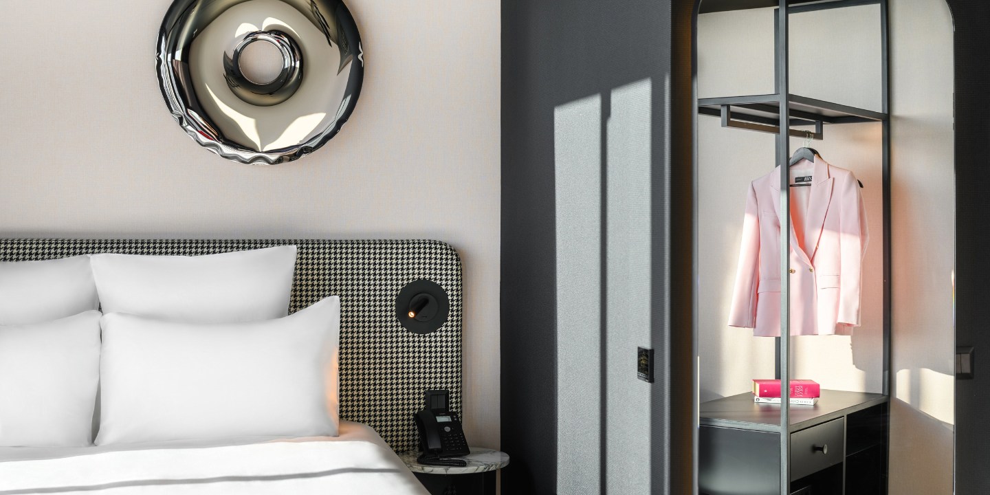 Une chambre d’hôtel élégante avec un lit moelleux aux oreillers blancs, un blazer rose suspendu dans une penderie moderne et un téléphone sur la table de chevet.