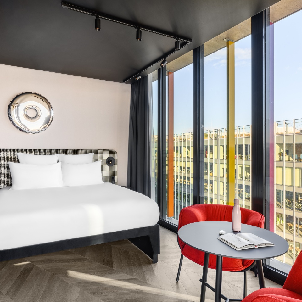 Une chambre d’hôtel avec un lit King size, des chaises rouge vif et de grandes fenêtres aux encadrements colorés baignées de lumière naturelle.
