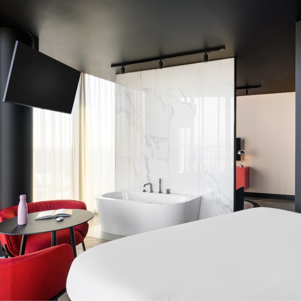 Chambre d’hôtel avec une élégante baignoire îlot, un mobilier épuré et de grandes fenêtres aux rideaux fluides. Coin salon douillet et télévision.