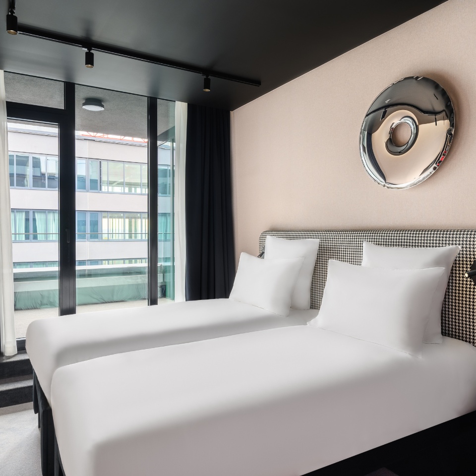 Chambre d’hôtel avec deux lits jumeaux soigneusement faits, de grandes fenêtres baignées de lumière naturelle et une décoration élégante.