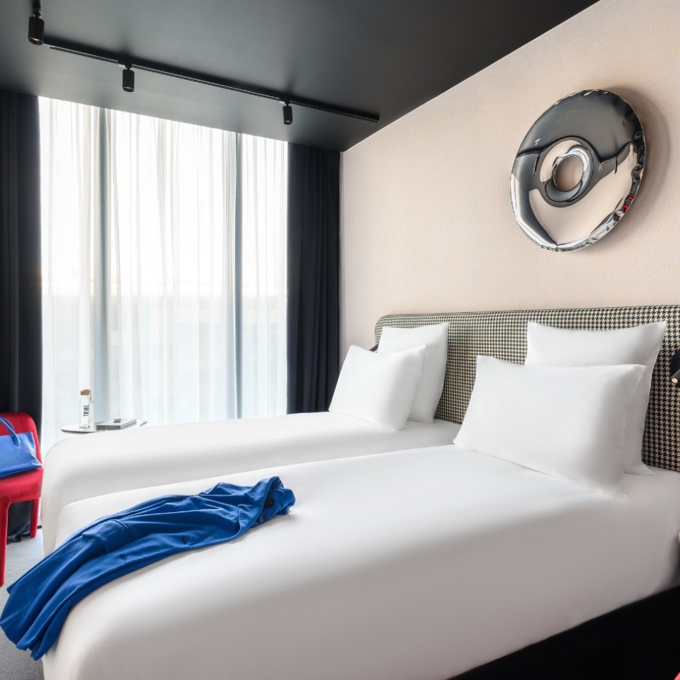 Chambre d’hôtel avec deux lits soigneusement faits, un vêtement bleu posé sur l’un d’eux et une décoration artistique en métal, la lumière filtrant à travers des voilages.