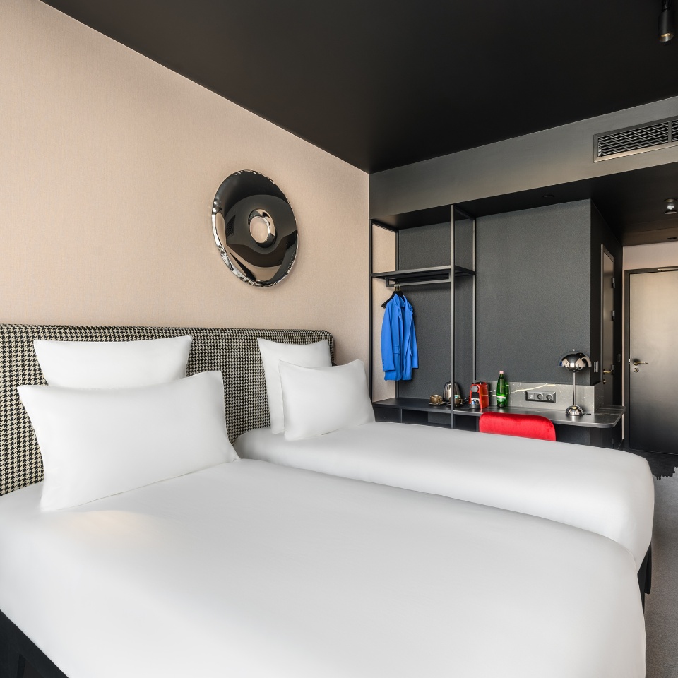 Chambre d’hôtel avec deux lits jumeaux, un miroir mural élégant et un bureau au design épuré, dans des tons neutres rehaussés de touches de couleur.