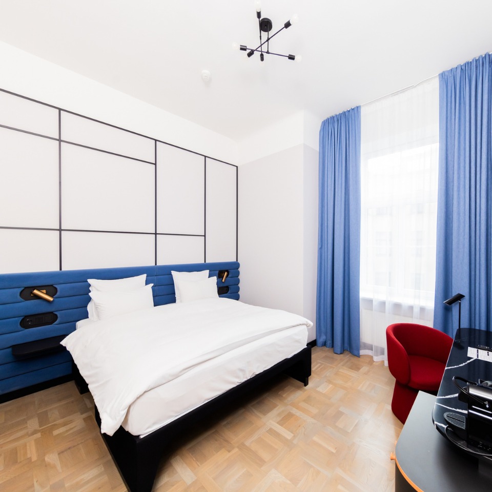 Modern hotel room featuring a blue upholstered bed, white linens, wooden floor, and a red chair by a desk with a window draped in blue curtains.