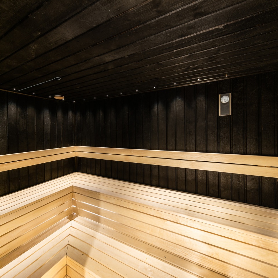 Interior of a modern sauna featuring black wooden walls and light-colored wooden benches, with subtle lighting and a thermometer.