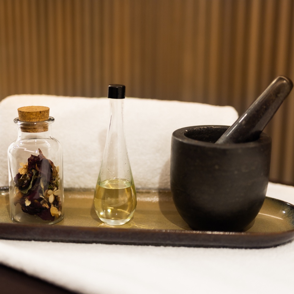 A tray holds a glass jar of dried herbs, a narrow bottle of oil, and a black mortar and pestle, set against soft towels.