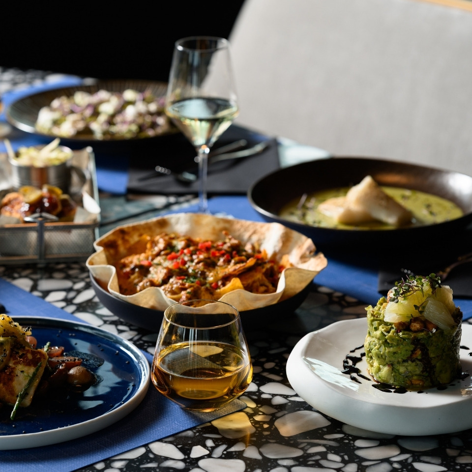 Varied gourmet dishes and drinks served on a marble table with blue placemats, featuring appetizers, entrees, and wine glasses.