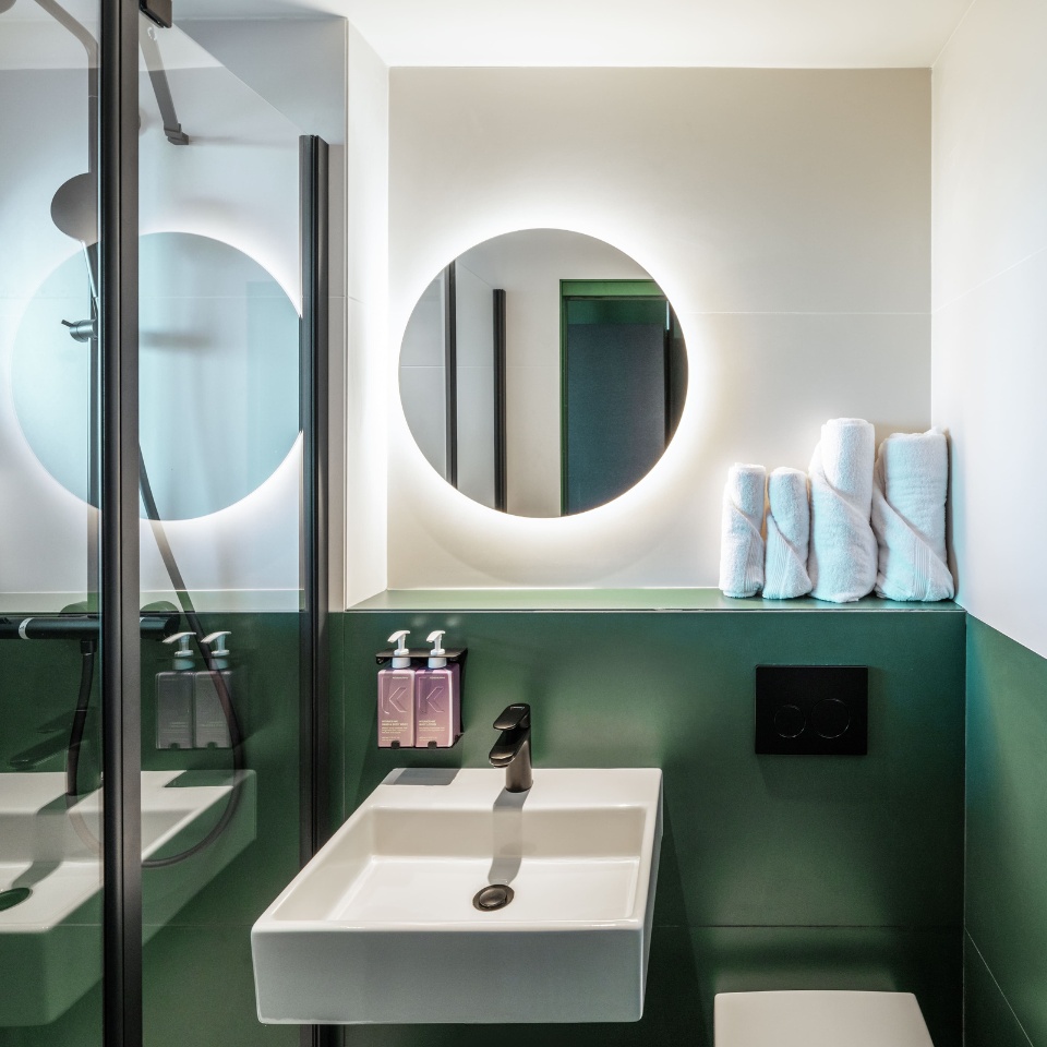 Bathroom with a glass shower, green tiles and a round mirror.