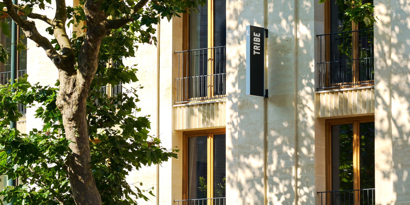 Hotel façade with balconies and a TRIBE sign behind trees.