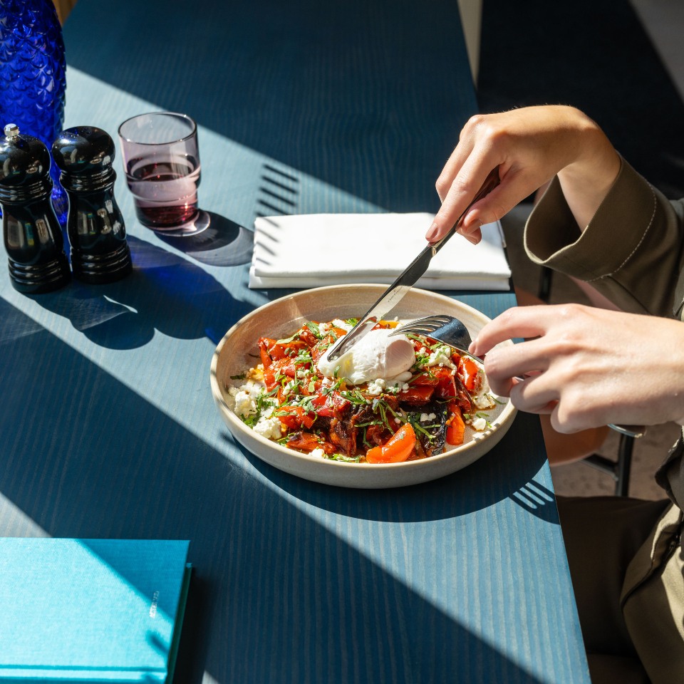 A hand holds a fork above a vibrant salad topped with poached egg, sitting on a sunlit table with condiments and a glass nearby.