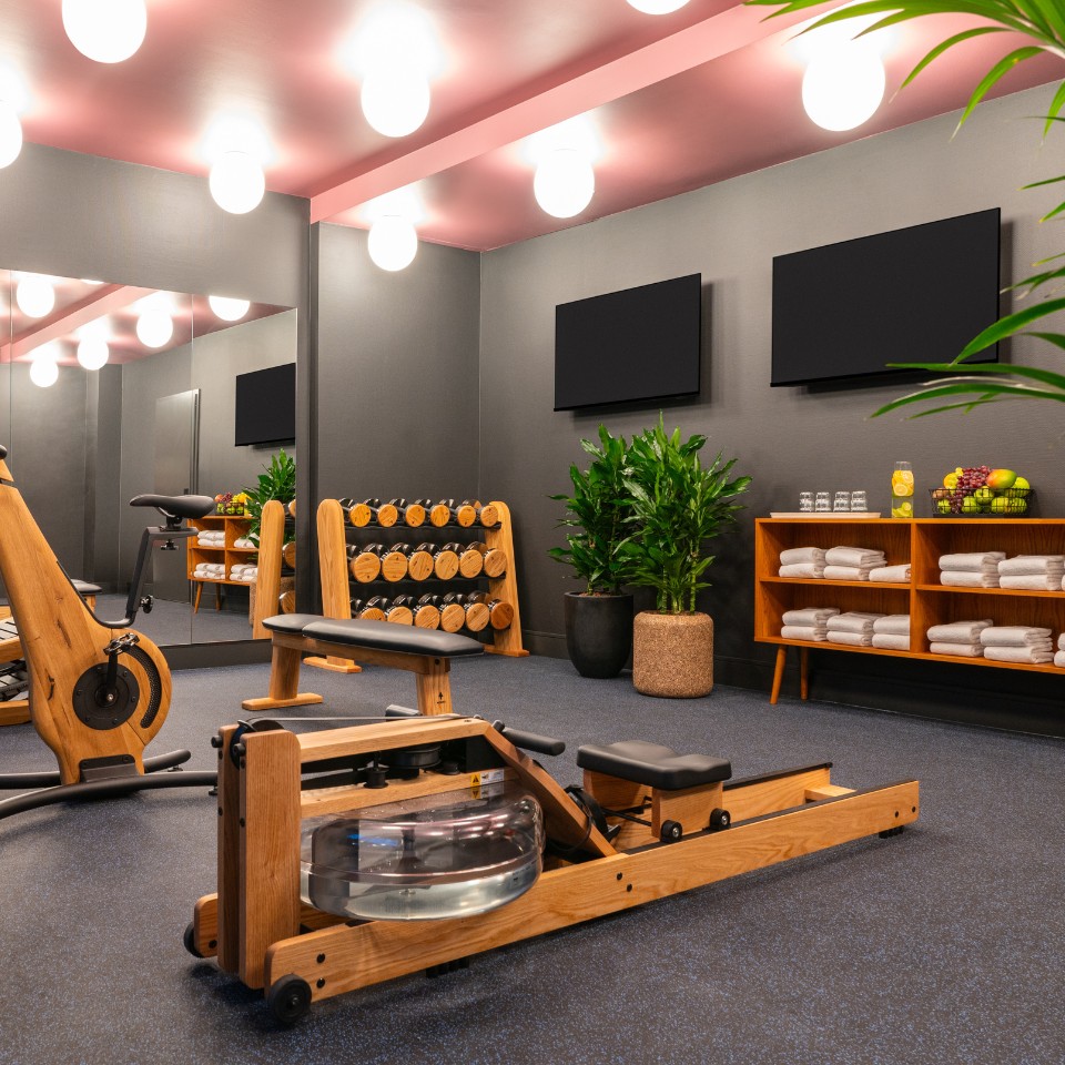 Modern fitness studio with wooden rowing machine, dumbbell rack, plants, and neatly arranged towels and refreshments under bright lights.