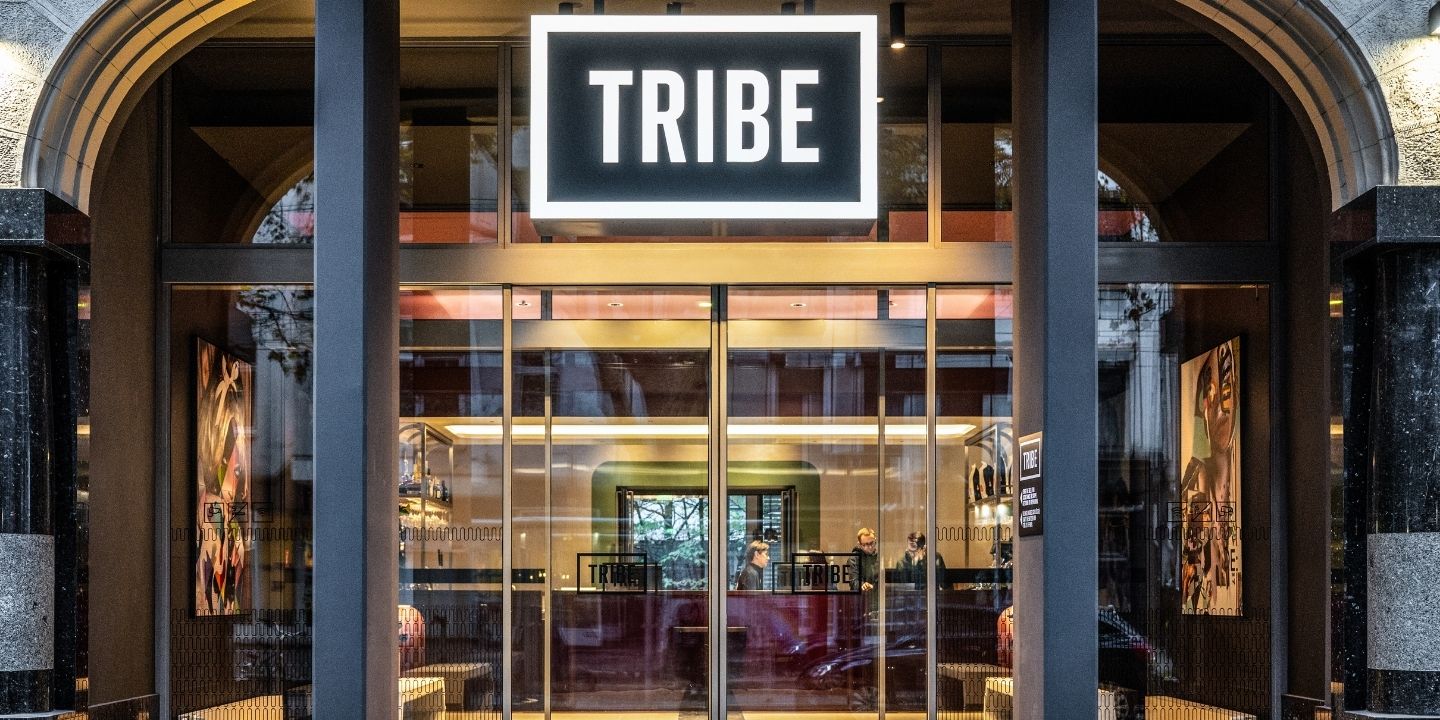 Modern hotel entrance labeled 'TRIBE,' showcasing glass doors and stylish interior, with staff engaging guests inside.
