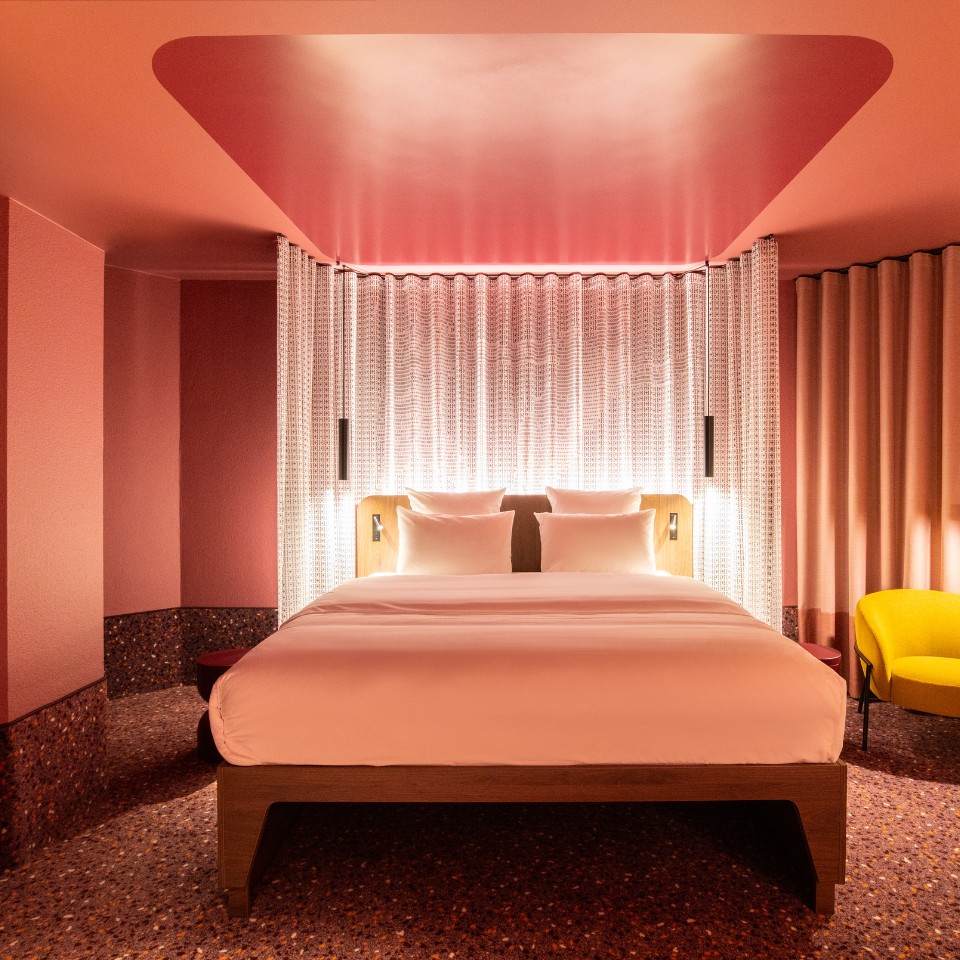 A hotel room featuring a pink color scheme, a bed with white pillows, decorative lighting, and a yellow accent chair.