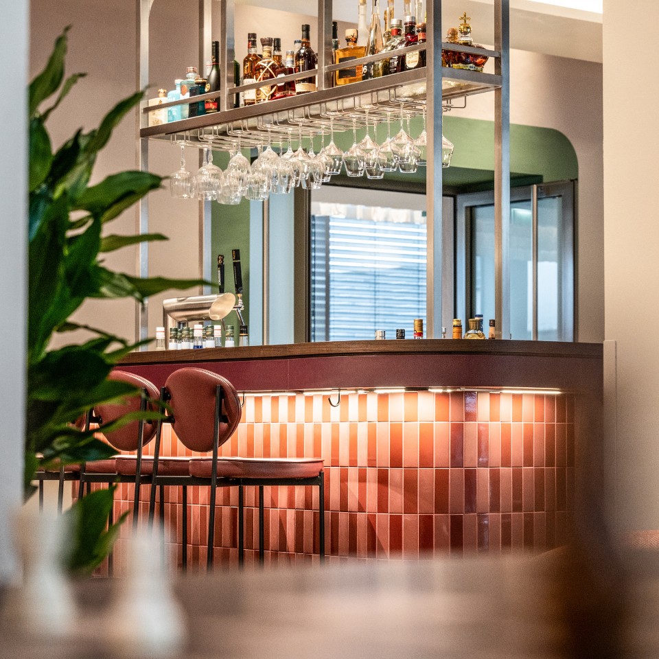 A cozy bar with two brown padded stools, lined with colorful bottles and glasses, framed by warm lighting and green plants.