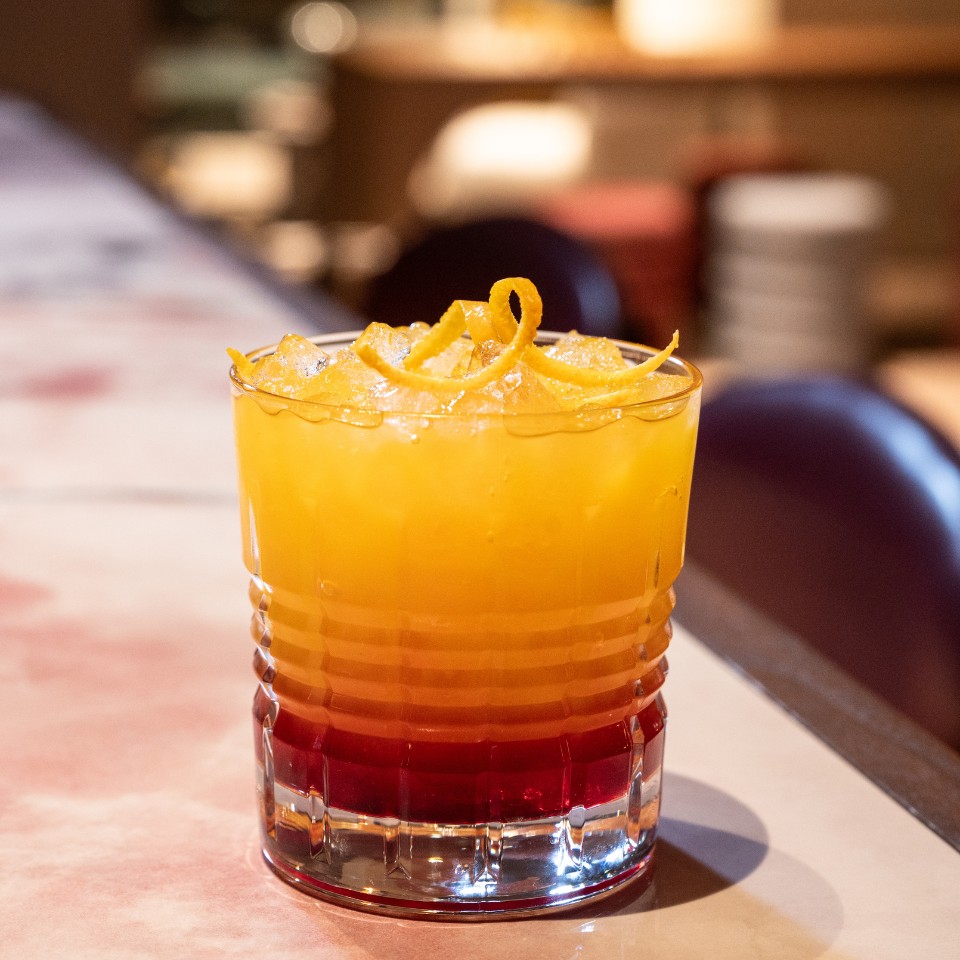 A vibrant cocktail layered with orange and red hues, garnished with citrus twists, served on a bar countertop.