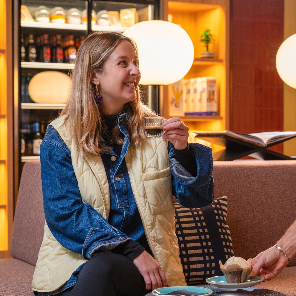 A woman in the hotel lobby holds an espresso while seated, with snacks and drinks on a table beside her. Soft lighting creates a warm atmosphere.