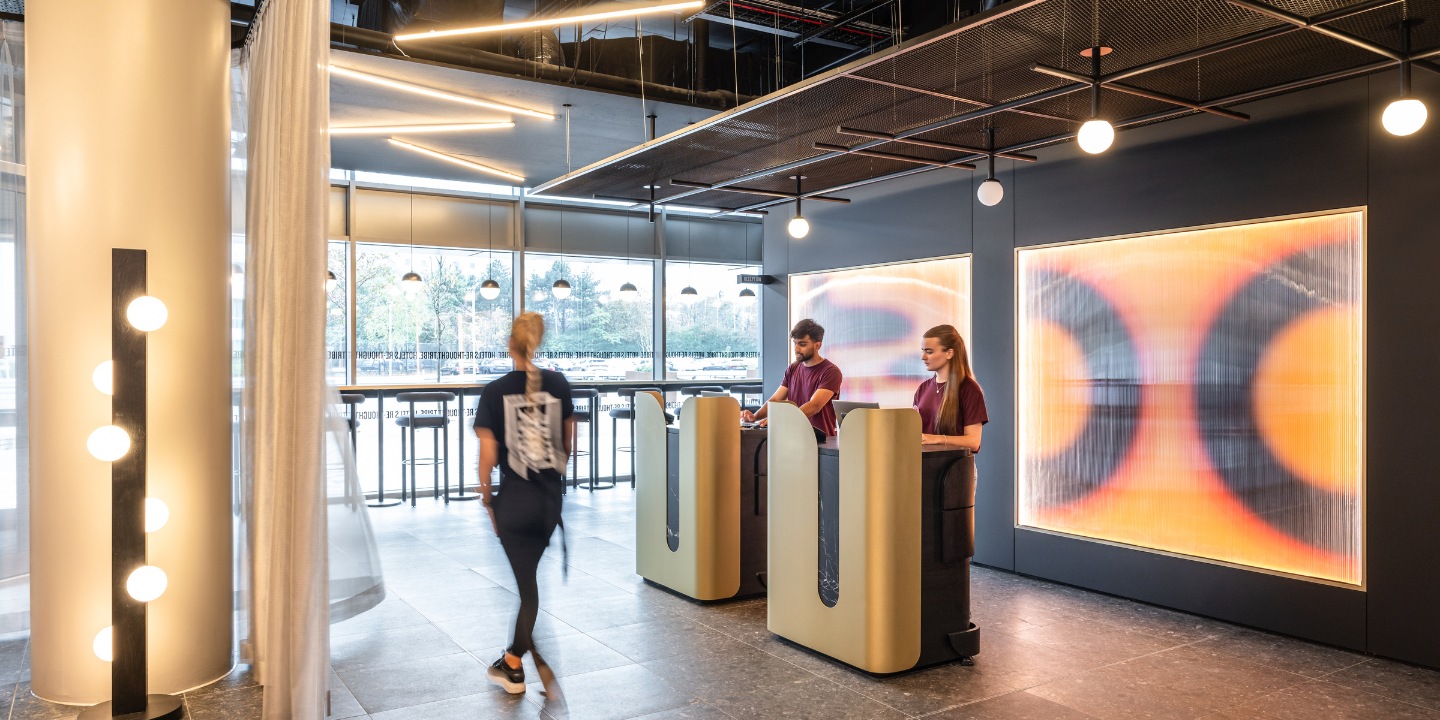 A modern reception area features two employees at stylish desks, with decorative lighting and an artistic backdrop.