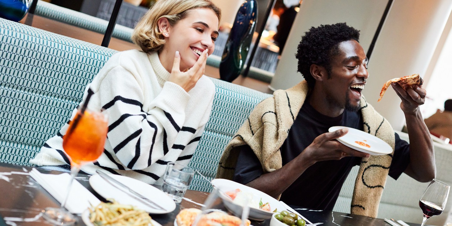 A dining table is filled with various dishes, including pizzas, pasta, and olives, as two people enjoy their meal in a cozy restaurant setting.