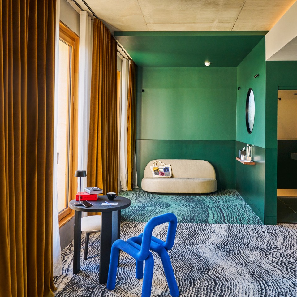 Coin salon aux murs verts, avec rideaux moutarde, fauteuil bleu et canapé beige près de la fenêtre.
