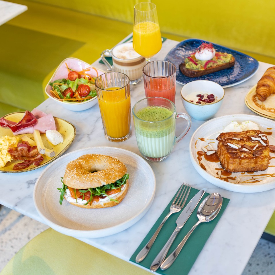 A vibrant breakfast spread featuring a bagel sandwich, assorted drinks, salads, croissants, and sweet dishes on a marble table.