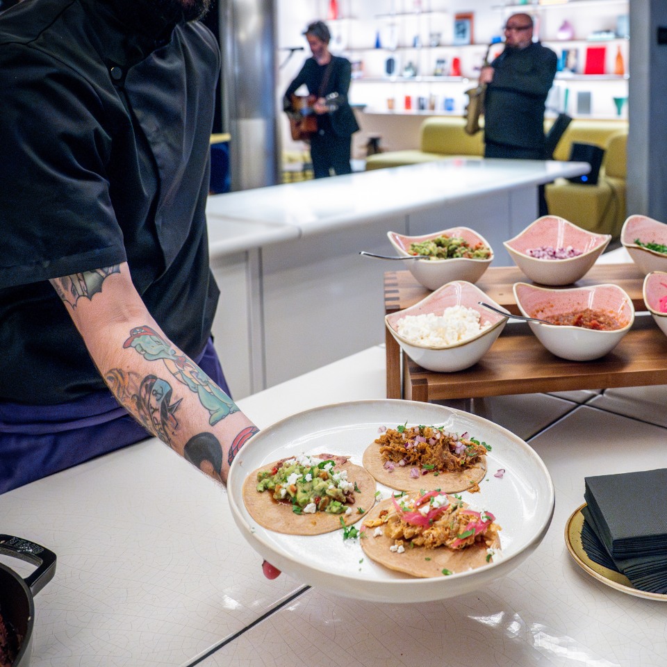 A chef presents a plate of three colorful tacos, garnished with fresh toppings, amidst a vibrant dining scene.