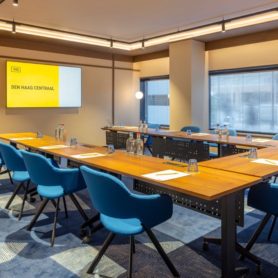 Modern conference room with wooden tables arranged in a U-shape, blue chairs, and bottles of water. A yellow presentation screen reads "Den Haag Centraal."