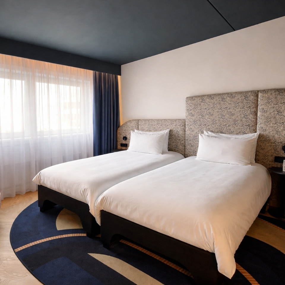 Modern hotel room featuring two twin beds, warm lighting, and sheer curtains, with a stylish rug and patterned headboard.