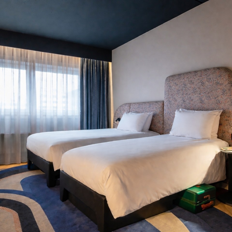 A modern hotel room featuring two twin beds with white linens, a large window with sheer curtains, and stylish decor on the walls.