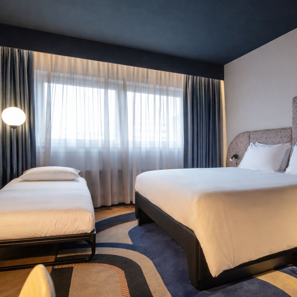 Modern hotel room with a large bed, single bed, sheer curtains, dark blue ceiling, and a round wall lamp.