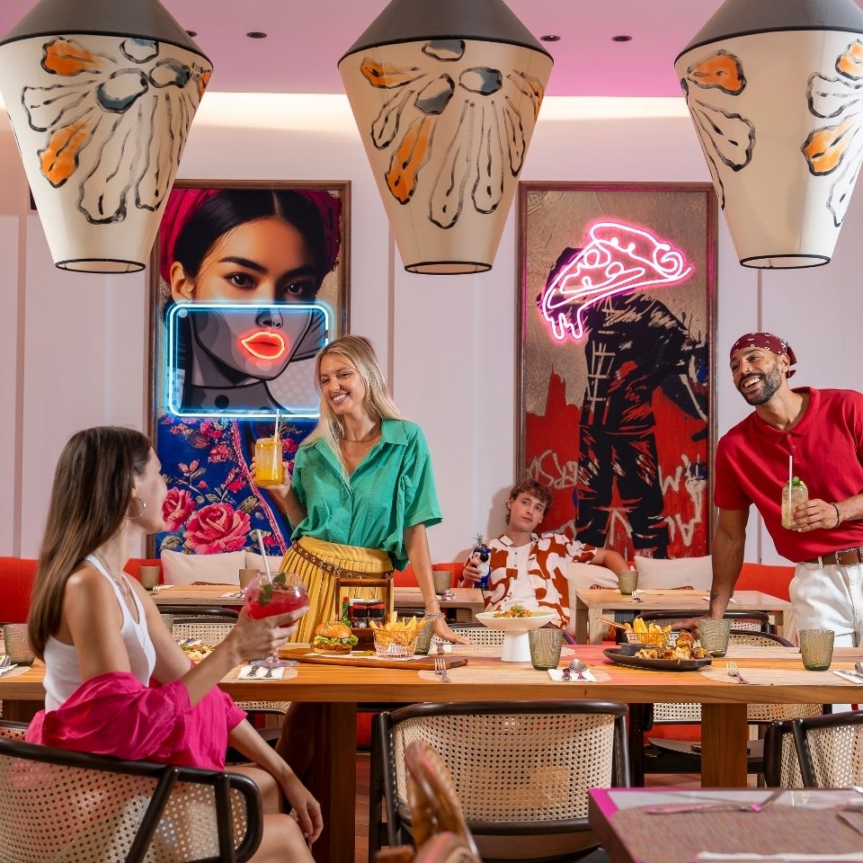 A vibrant restaurant scene with three friends laughing around a table filled with food. Neon lights and colorful art create a lively, fun atmosphere.