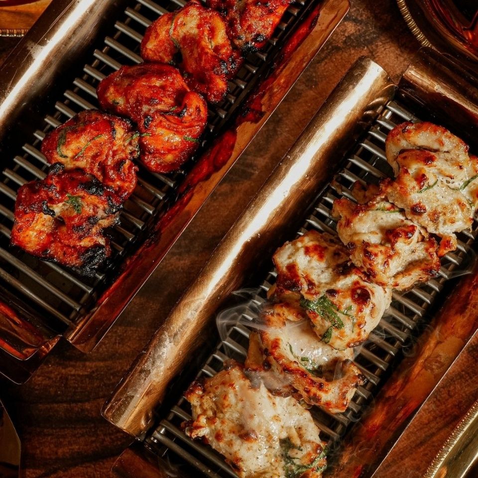 Deux sortes de brochettes de poulet mariné grillé, l’une rougeâtre et l’autre blanche, servies sur des grilles rectangulaires en métal.