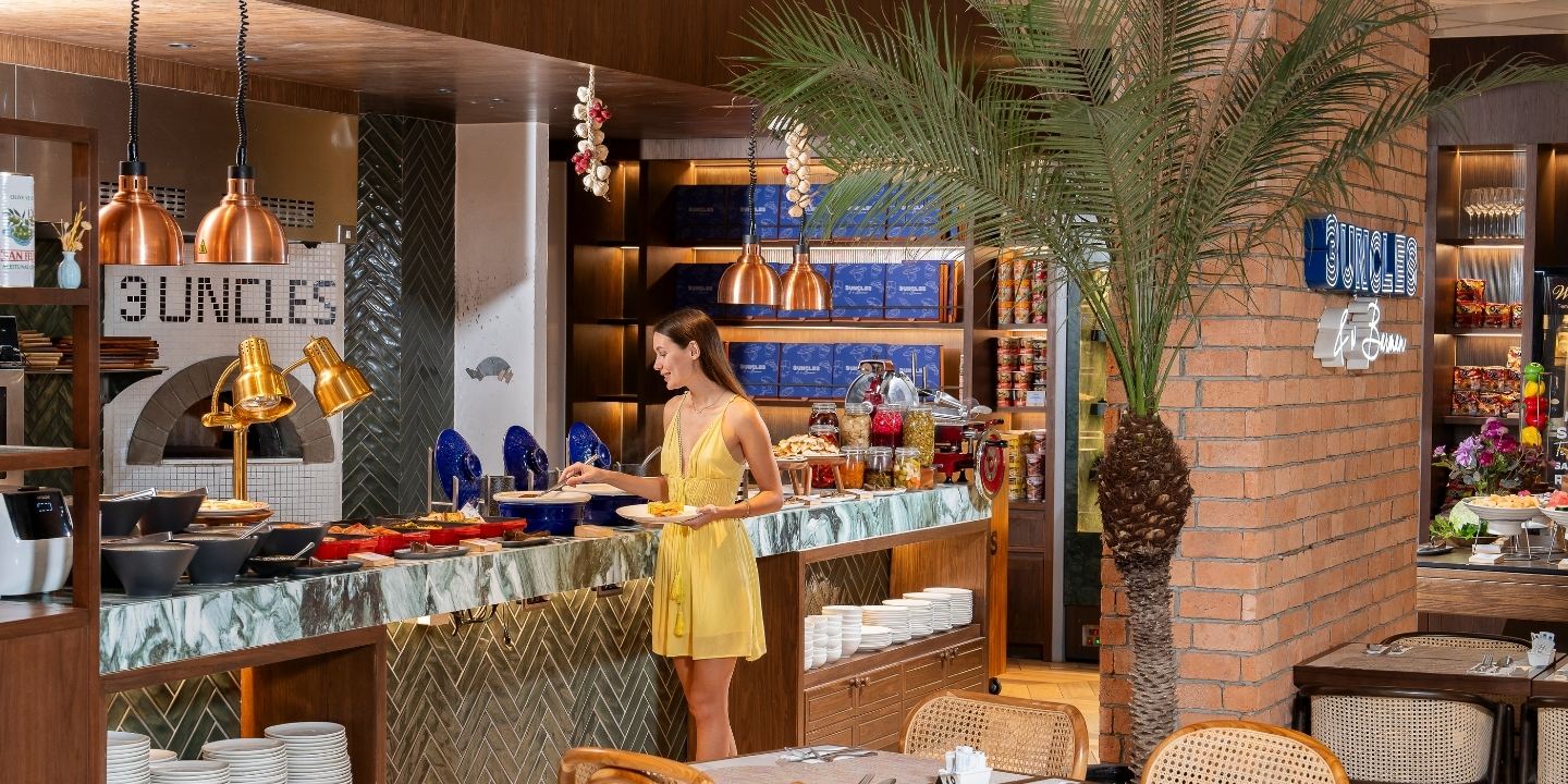A woman in a yellow dress selects food from a buffet in a stylish restaurant. The setting features warm lighting, wooden decor, and a palm plant, creating a cozy ambiance.