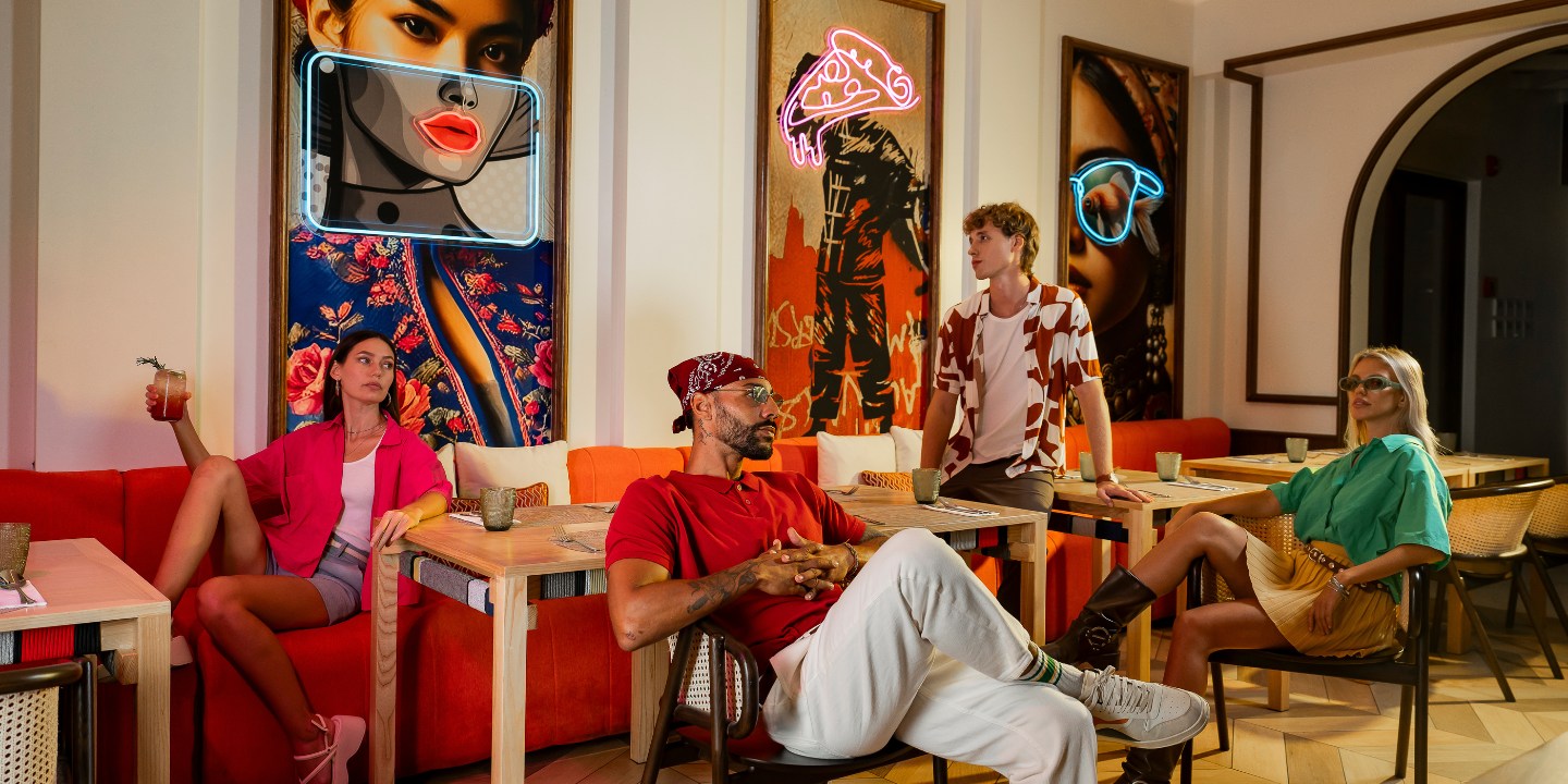 Four people relax in a vibrant, art-filled cafe with neon lights. There's a relaxed, stylish atmosphere, with pop art portraits on the walls.