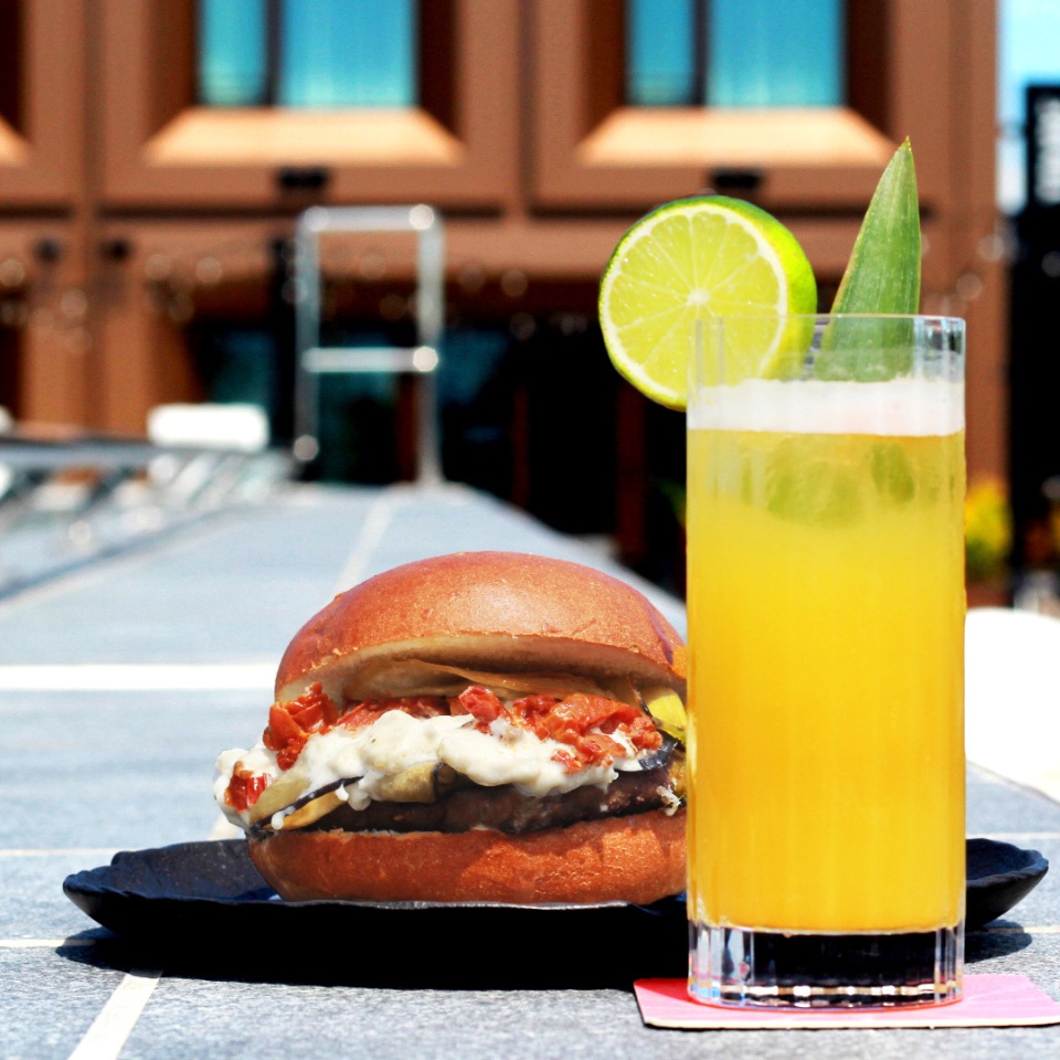 A juicy burger with toppings sits on a black plate next to a tall glass of orange drink garnished with lime, set on a sunny table.