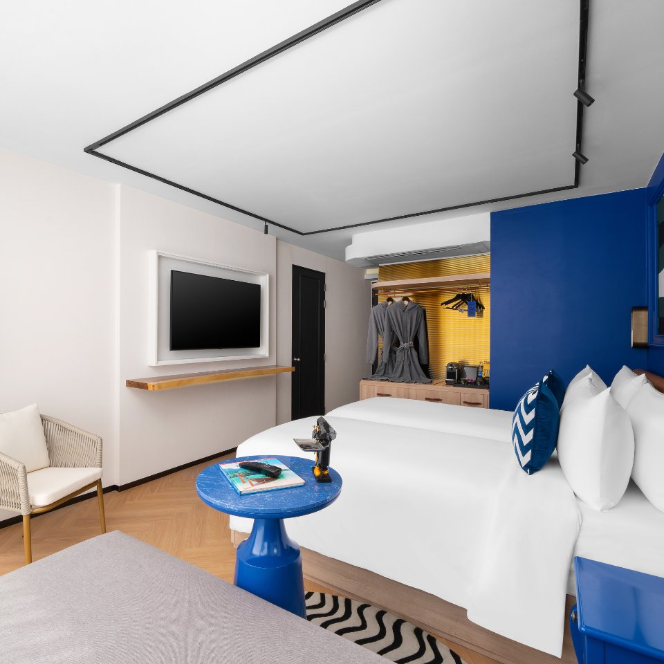 Bright hotel room featuring a double bed, blue accent wall, TV, and modern furnishings, including a small blue side table.