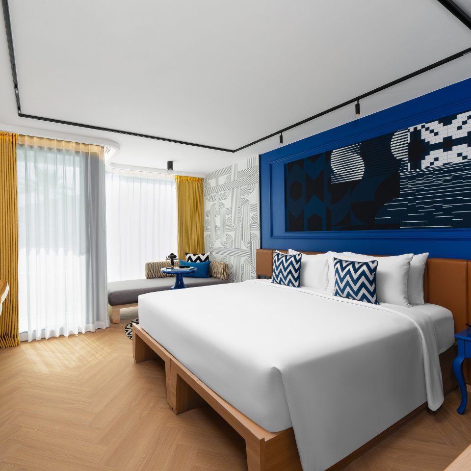 Modern hotel room with a large white bed, chevron-patterned pillows, vibrant blue accents, abstract art, and natural light from large windows with sheer curtains.