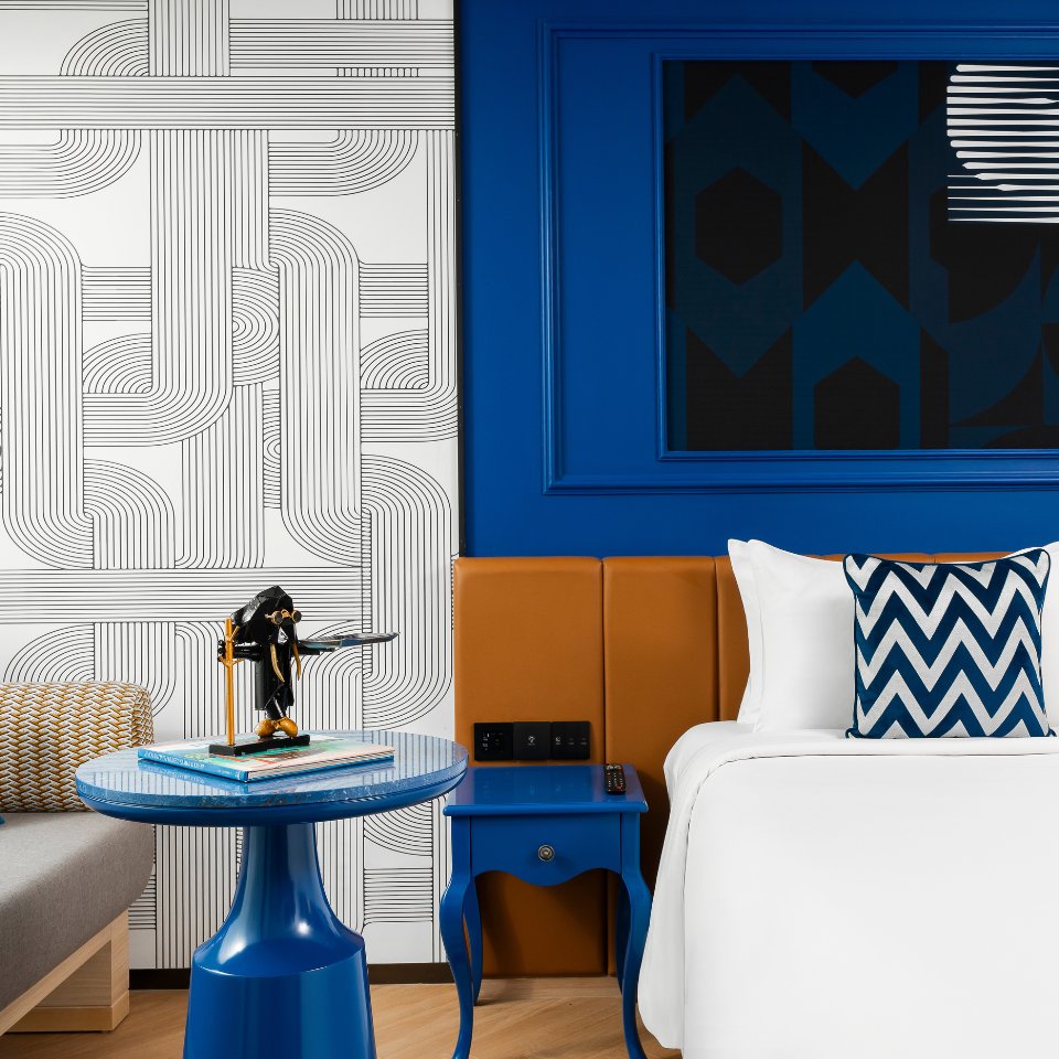 Modern hotel room with geometric designs. Features a white bed with a chevron pillow, blue accents, and patterned walls creating a stylish, vibrant feel.