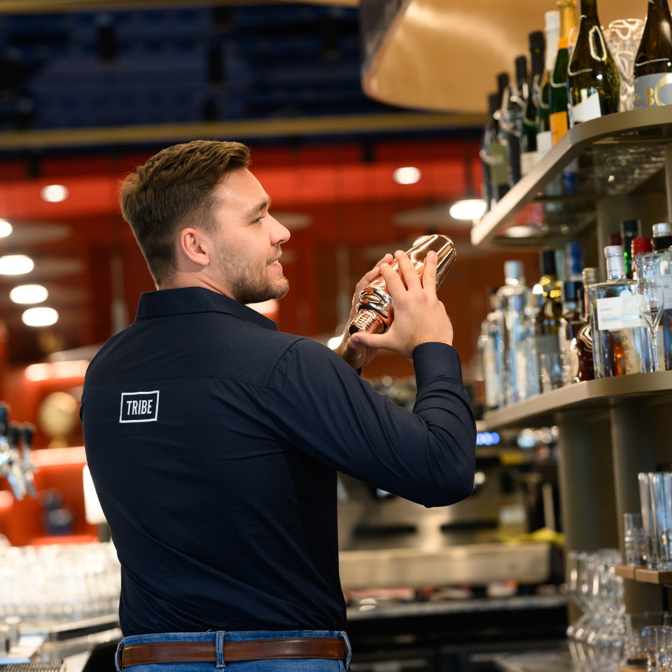 Bartender shaking a cocktail with a smile at a modern bar. Shelves filled with bottles in the background, conveying a lively, welcoming atmosphere.