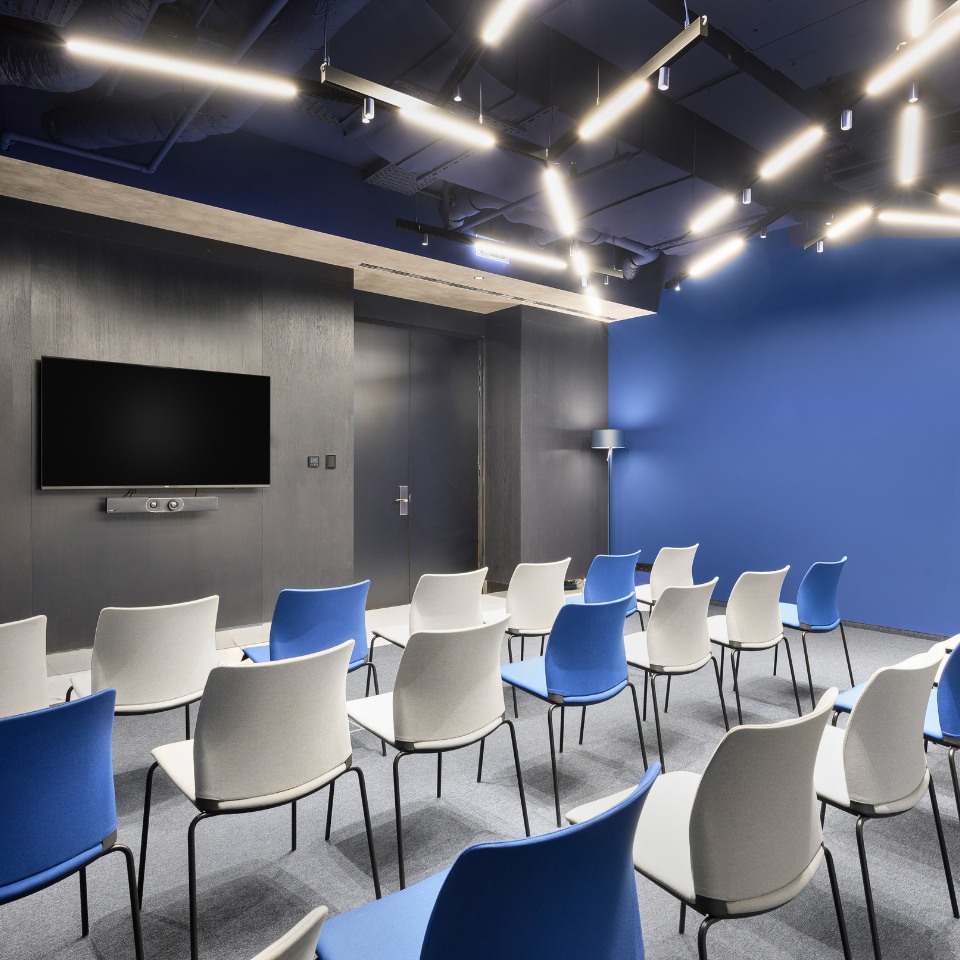 Modern conference room with blue and white chairs arranged in rows, a large screen on a dark wall, and bright overhead lights creating an inviting ambiance.