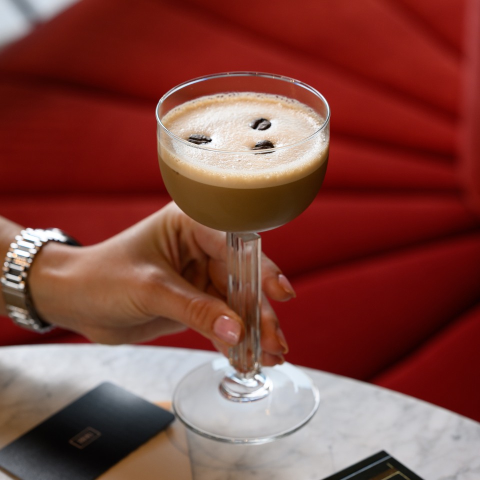 A hand holds an espresso martini with coffee beans on the frothy top. The background features a red cushioned seat, creating an elegant, cozy atmosphere.