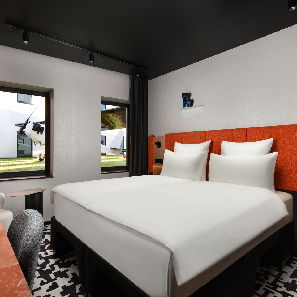 Modern hotel room with a neatly made bed featuring white linens and an orange headboard. Large windows show an airplane outside, giving a unique view.