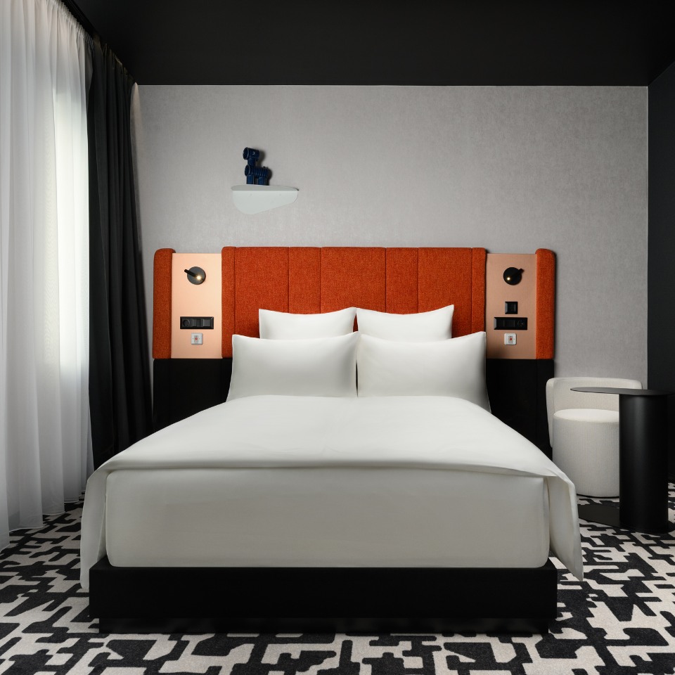 Modern hotel room with a plush bed featuring white linens, bold orange headboard, black-and-white patterned carpet, and sleek minimalist decor.