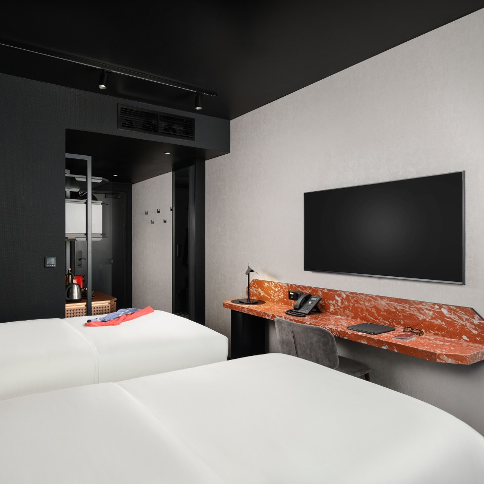 Modern hotel room with two white beds, a sleek black ceiling, and a glossy red marble desk with a lamp, phone, and TV. Minimalist and inviting atmosphere.