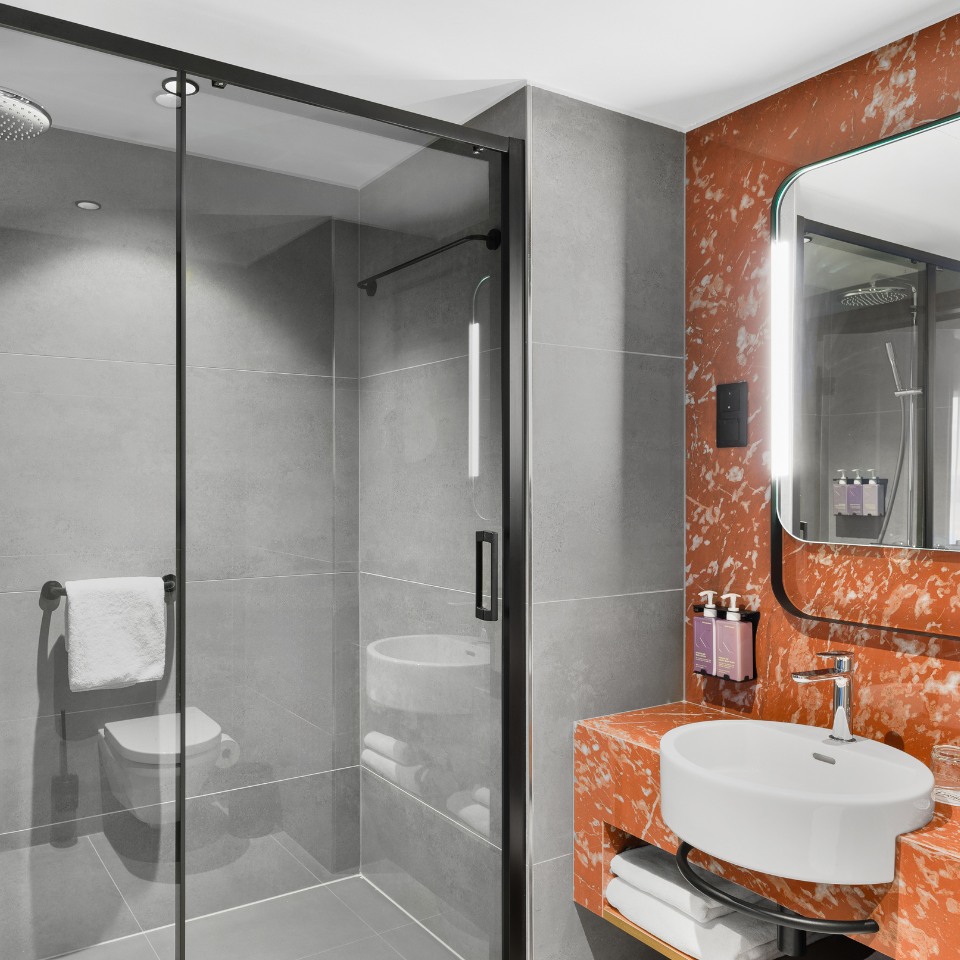 Modern bathroom with gray tiled walls and a walk-in shower with glass door. Features a marble-textured orange sink area and a rectangular backlit mirror.