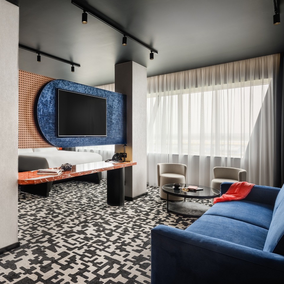 A modern hotel room with a geometric carpet, blue sofa, and large TV mounted on a stylish wall. White curtains let in soft light, creating a cozy atmosphere.