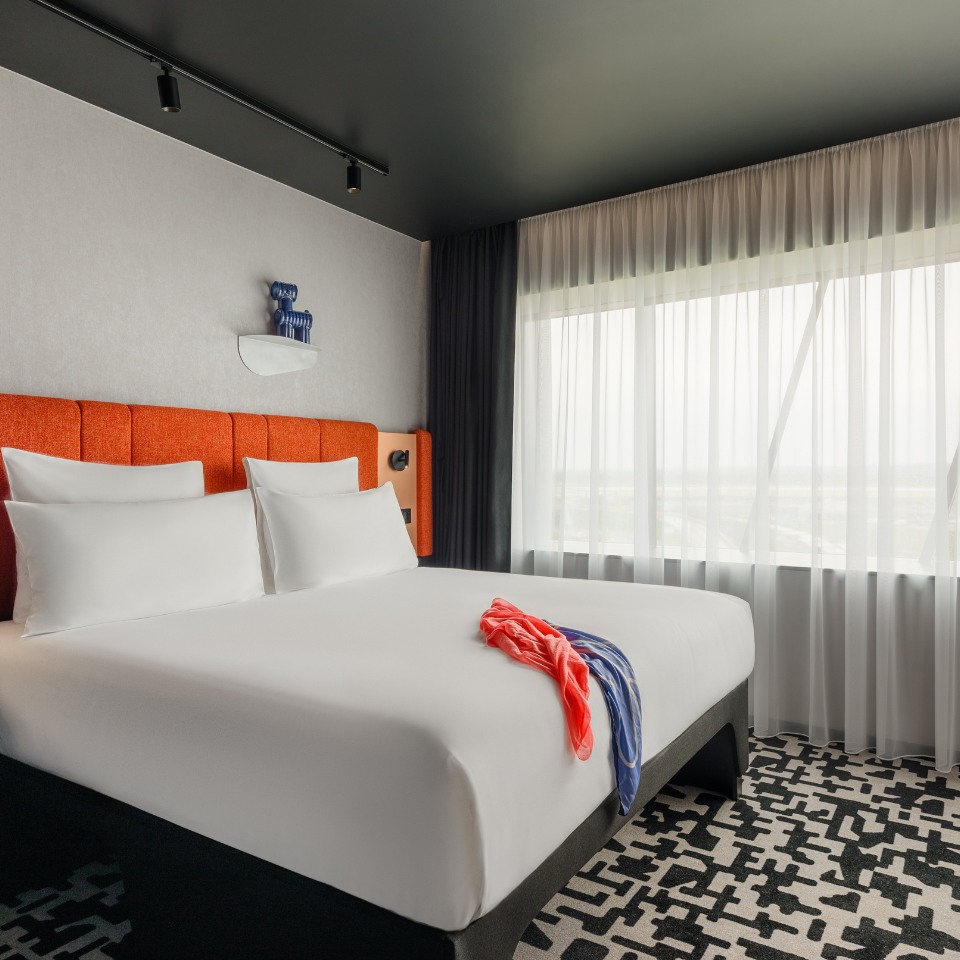 Modern hotel room with a cozy bed featuring an orange headboard, white linens, and colorful clothes. Large window with sheer curtains, geometric carpet. Tranquil ambiance.