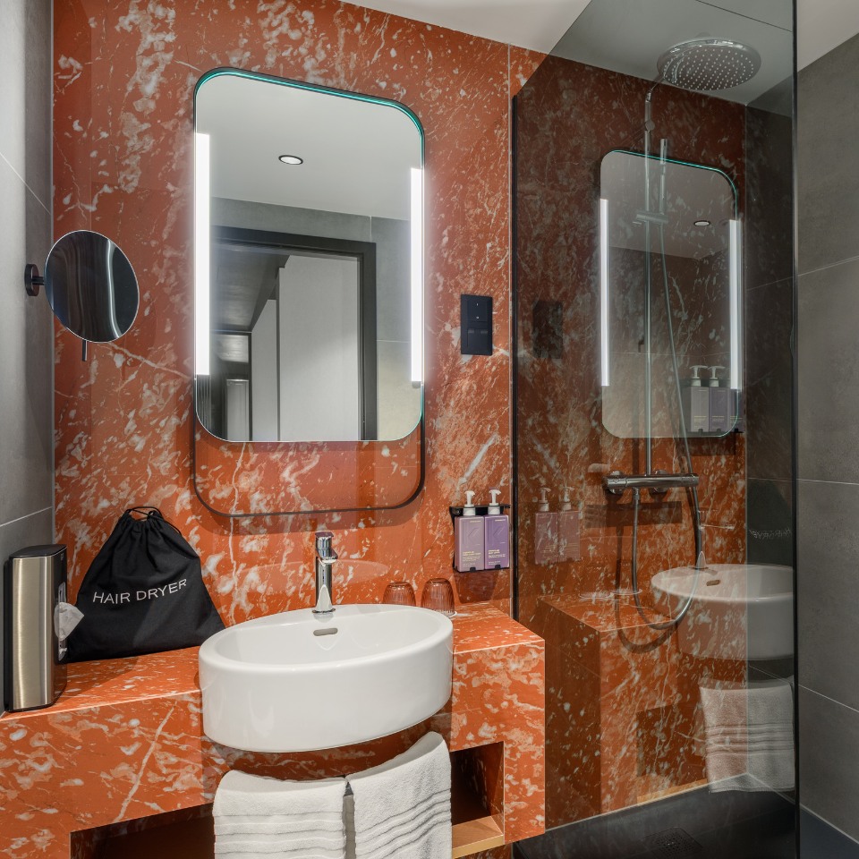Modern bathroom with orange marble walls, illuminated mirror, and sleek white basin. Glass shower cubicle visible. Chic, clean, and stylish ambiance.