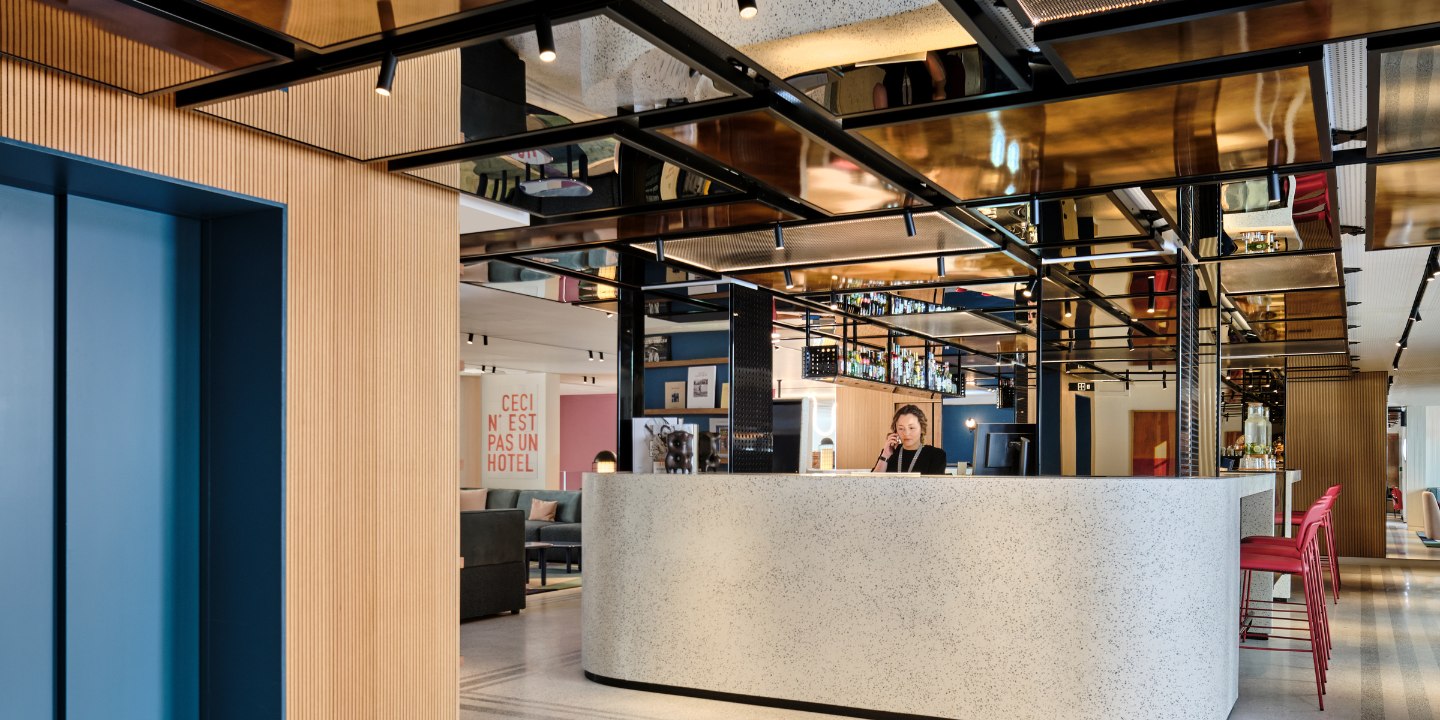 Modern hotel lobby with reflective ceiling, a sleek white reception desk, and a person on the phone. Art on the walls creates a chic, welcoming vibe.